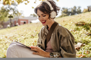 lovely-woman-jacket-headphones-sitting-grass-outdoors-happy-woman-with-curly-hairstyle-holding-smartphone-outside Medium